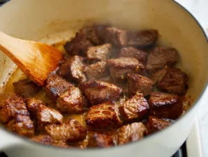 Browning beef cubes in a white Dutch oven with a wooden spoon